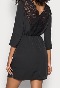 Schwarzes Kleid mit einem Spitzenrücken, dreiviertellangen Ärmeln und einer taillierten Passform mit seitlichen Schlitzen für zusätzlichen Stil. Glatte, weiche Textur.