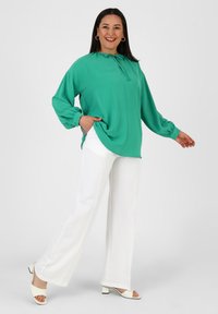 Grüne Bluse mit langen Ärmeln und Schleifenkragen, kombiniert mit weißen weit geschnittenen Hosen und weißen Blockabsatzsandalen. Einfaches Design und glatte Textur.