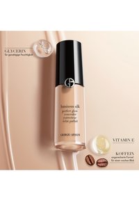 Flacon du correcteur Luminous Silk avec un capuchon noir, présenté avec une goutte de glycérol, une capsule de vitamine E et des grains de café sur un fond de fond de teint beige.