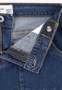 Denim jeans v temno modri barvi, ki imajo oranžne šive in kovinsko zadrgo. Vključujejo etiketo z oznako "Redna" in "Narejeno v Bangladešu."