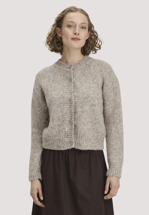Frau mit lockigem Haar, die eine beige gestrickte, geknöpfte Strickjacke und einen dunklen Rock trägt, steht vor einem schlichten Hintergrund.