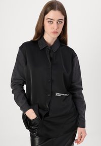 Melna satīna krekls ar džinsa apkakli un piedurknēm, ar kabatu un izšūtu "Karl Lagerfeld Jeans" logotipu priekšpusē.