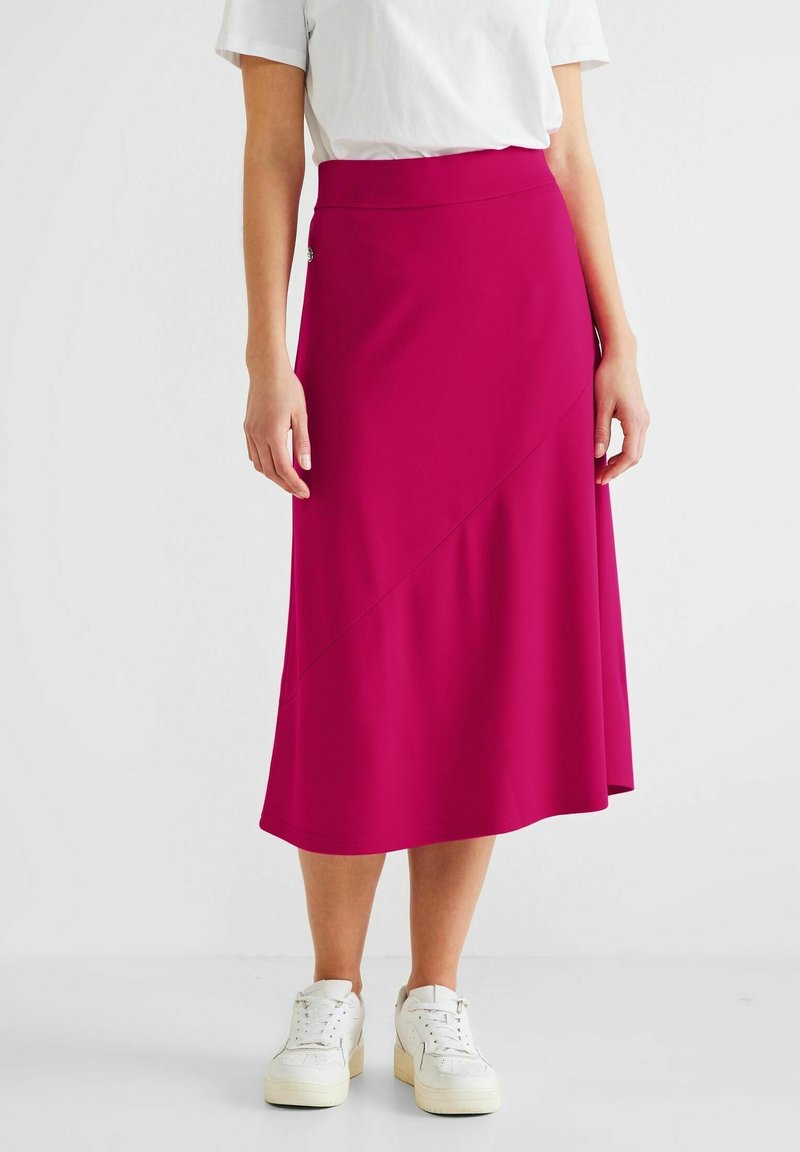 Femme portant une jupe midi rose vif avec une couture diagonale, un t-shirt blanc et des baskets blanches, debout sur un fond uni.