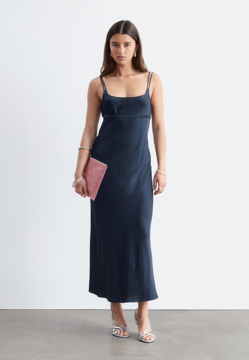 Femme portant une robe maxi bleu marine sans manches, tenant une pochette rose, des bracelets argentés, des boucles d'oreilles et des talons hauts argentés à brides.