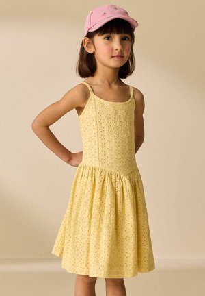 Girl with straight dark hair wearing a pink cap and sleeveless yellow eyelet dress, standing with hands on hips against beige background.