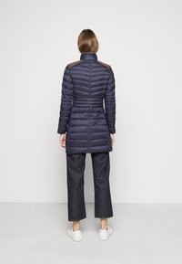 Marineblauer, gesteppter längerer Mantel mit hohem Kragen, braunen Schulterakzenten, tailliertem Schnitt und kombiniert mit dunklen, weit geschnittenen Jeans und weißen Sneakers.