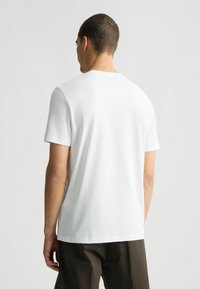 Tricou alb din bumbac, cu mâneci scurte, guler rotund, textură netedă, vedere din spate arată un design simplu cu un croi drept la tiv.