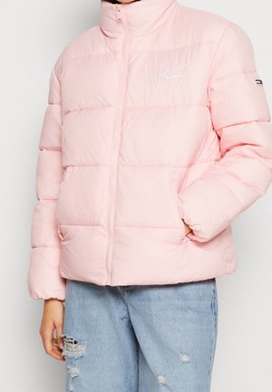 Personne portant une veste matelassée rose clair avec une fermeture éclair sur le devant et un logo brodé sur la poitrine, associée à un jean bleu clair déchiré.