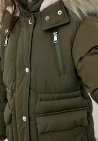 Veste matelassée vert foncé avec un col en fausse fourrure, dotée de boutons-pression argentés et de poches zippées. Texture douce et matelassée avec des coutures apparentes.