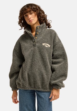 Femme aux cheveux bouclés portant un pull polaire gris à boutons avec le logo Billabong, associé à un jean bleu, debout devant un fond blanc.
