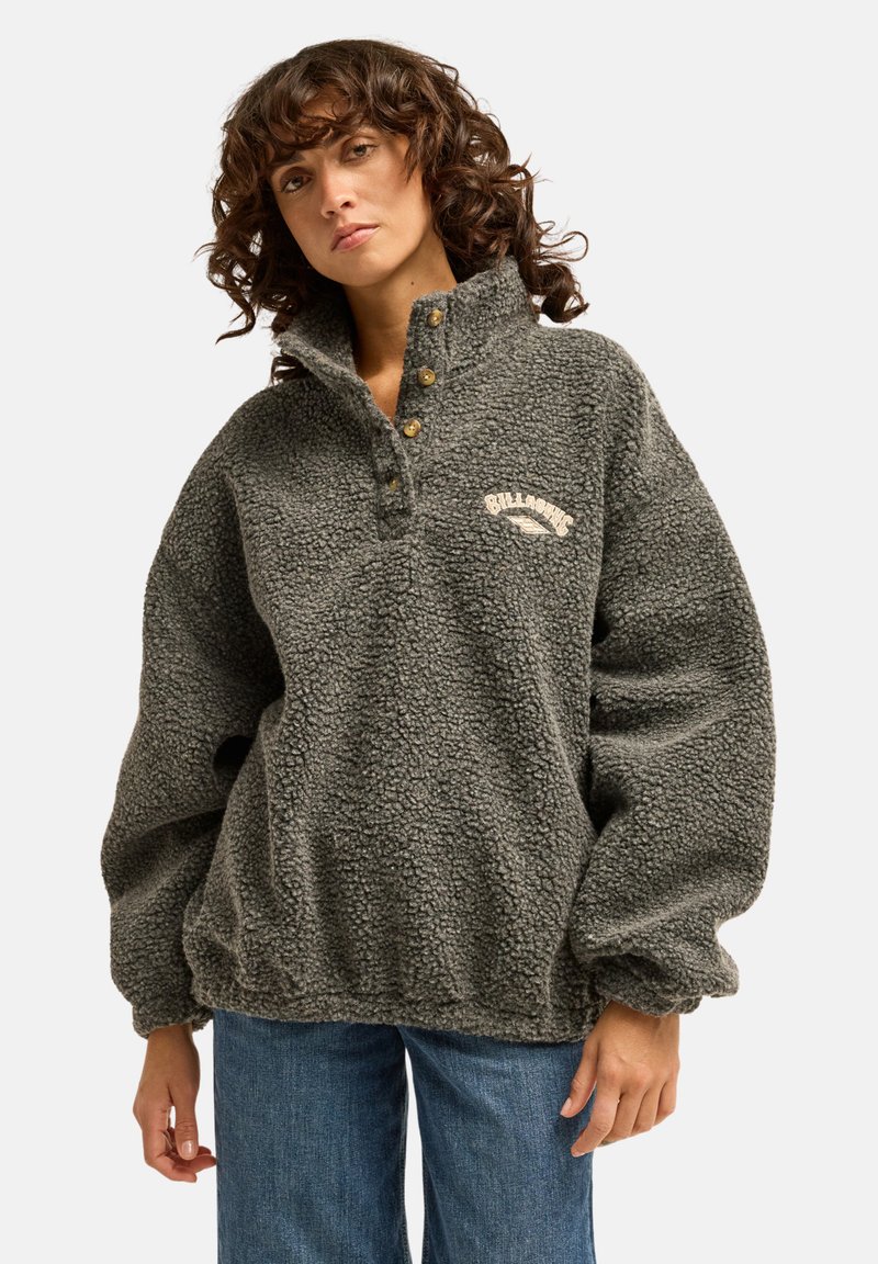 Femme aux cheveux bouclés portant un pull polaire gris à boutons avec le logo Billabong, associé à un jean bleu, debout devant un fond blanc.