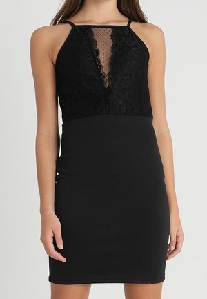 Kvinde iført en ærmeløs sort kjole med blonde- og gennemsigtigt stofdetalje øverst på brystet og en tætsiddende, ensfarvet sort nederdel.