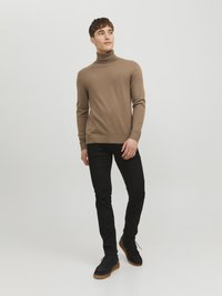 Beige Rollkragenpullover, eng anliegend, leichtes Strickmaterial. Kombiniert mit schwarzen Slim-Fit-Jeans und dunklen Wildlederschuhen mit Gummisohlen.