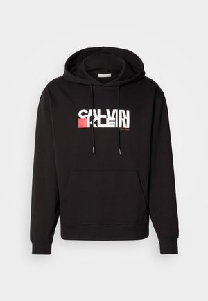 Melnā kapuce ar priekšējo kabatu, auklu kapuci un balto/sarkano logotipu. Izgatavota no mīkstas auduma ar ribotām manšetēm un apakšmalu.