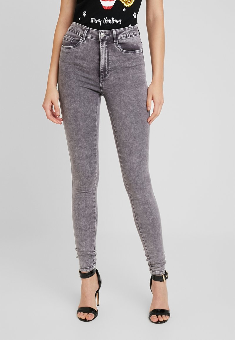 Jeans skinny taille haute gris avec une texture délavée et des détails de couture, associés à un t-shirt graphique noir et des sandales à talons noires.