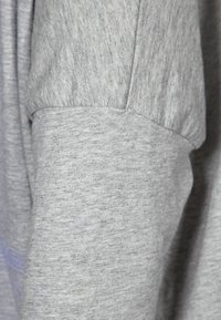 Gros plan d’un tissu gris avec une couture visible et un détail de poignet, montrant une texture tricotée douce et chinée.