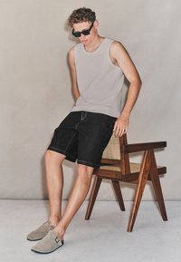 Lichtgrijze, mouwloze tanktop, zwarte denim shorts, grijze suède instapschoenen. Model zittend op een houten stoel met een gevlochten rugleuning.