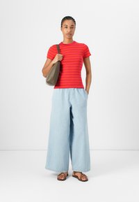 T-shirt rayé rouge, large pantalon en denim bleu clair, sandales à brides marron, tenant un sac gris, se tenant devant un fond blanc uni.