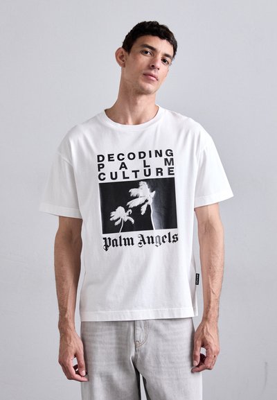 T-shirt en coton blanc avec un design graphique comportant le texte "DÉCODAGE DE LA CULTURE PALMIER", une illustration de palmiers et le logo "Palm Angels" en noir.
