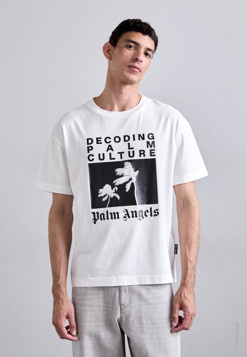 T-shirt en coton blanc avec un design graphique comportant le texte "DÉCODAGE DE LA CULTURE PALMIER", une illustration de palmiers et le logo "Palm Angels" en noir.