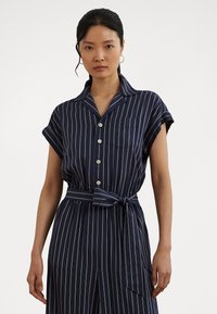 Marineblauw gestreepte shirtjurk met korte mouwen, knopen aan de voorkant en borstzak. Inclusief een gestrikt tailleband en lichtgewicht stof.
