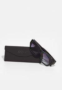 Black sunglasses with a geometric design feature gradient lenses, sitting beside a black, angular case embossed with "QUAY."