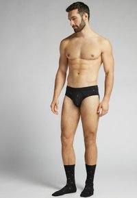 Czarne slipki w czerwony wzór kropkowy, zestawione z czarnymi skarpetkami w podobne kropki. Model stoi, prezentując dopasowanie i szczegóły wzornictwa.