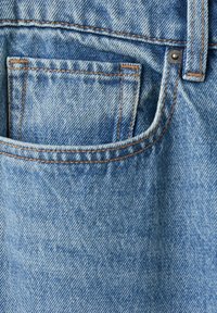Gros plan d'une poche de jean en denim bleu avec des surpiqûres orange et un rivet métallique près de la passants de ceinture.