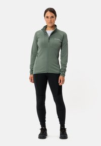 Veste en polaire zippée verte avec une surface texturée, associée à un legging noir. La veste présente un col haut et des poches latérales.