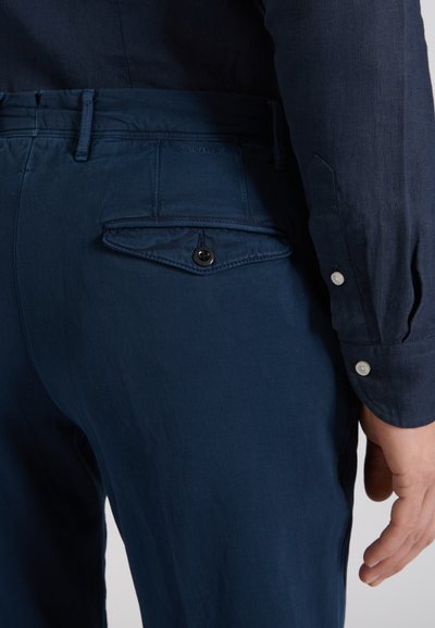 Close-up of person wearing dark blue pants with back buttoned pocket and matching dark blue long-sleeve shirt.