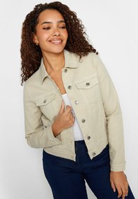 Beige denim takki, jossa on vartalonmyötäinen muotoilu, etuosassa napit ja kaksi rintataskua. Mallilla on se päällään valkoisen yläosan kanssa.