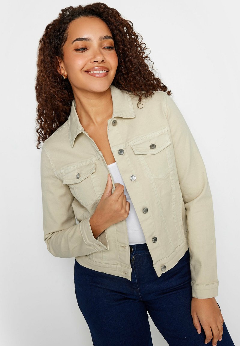 Beige denim takki, jossa on vartalonmyötäinen muotoilu, etuosassa napit ja kaksi rintataskua. Mallilla on se päällään valkoisen yläosan kanssa.