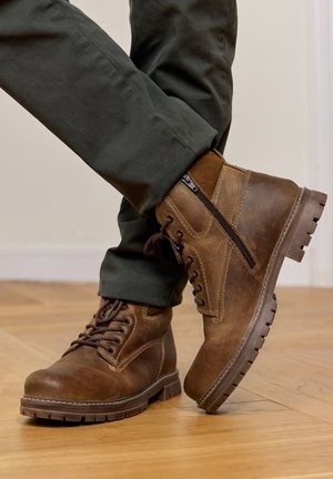 Braune Lederstiefel mit seitlichen Reißverschlüssen und Schnürsenkeln, getragen mit dunkelgrünen Hosen, stehend auf einem Holzboden, wobei ein Fuß über den anderen geschlagen ist.