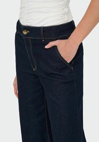 Mörkblå denimbyxor med avslappnad passform, utrustade med en guldfärgad knappstängning, sidofickor och kontrasterande guldstickning längs sömmarna.