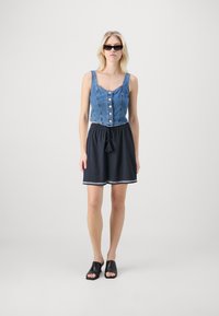 Džínový crop top s gombíkmi, skombinovaný s tmavomodrou sukňou so šnúrkou v páse a textúrovaným lemom, doplnený čiernymi sandálmi na otvorenej špičke.