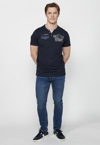 Tamno plava majica kratkih rukava s henley ovratnikom, džepom na prsima i natpisom "NORTH STAR." Uparena s svijetloplavim slim-fit trapericama i tamnim tenisicama.