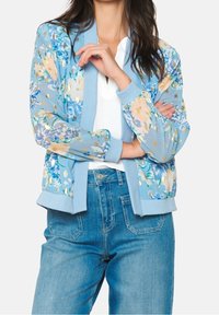 Světle modrá bomber bunda s květinovými vzory v růžové, bílé a zlaté akcenty. Žebrované manžety a lem, spárováno s vysokými džíny.