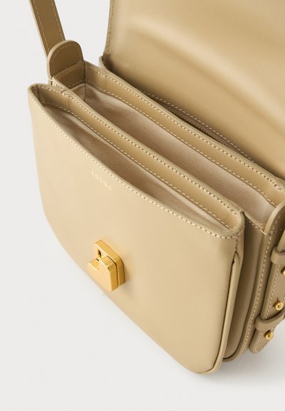 Μπεζ δερμάτινη τσάντα χιαστί με χρυσό κούμπωμα, δύο ανοιχτές θήκες και ρυθμιζόμενο λουράκι με χρυσά τρουκς σε λευκό φόντο.