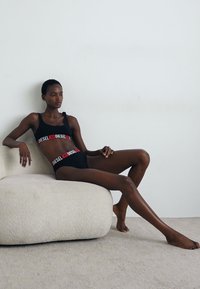 Bralette noire et culotte assortie avec un logo rouge "DIESEL" audacieux. Modèle assis sur une chaise crème douce et arrondie contre un fond uni.