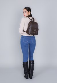 Mochila de cuero marrón con puntos dorados, tapa frontal con hebilla, dos correas para los hombros. El modelo lleva pantalones ajustados de mezclilla azul y botas negras hasta la rodilla.