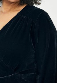 Melnā samta apakšveļa ar garām piedurknēm, kurai ir plisēta apdare uz pleciem un gluda tekstūra. Vienkāršs un elegants dizains.