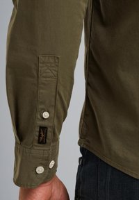 Camisa de manga larga verde oliva con puños abotonados, botones claros en contraste y detalle de bolsillo cosido con una etiqueta.
