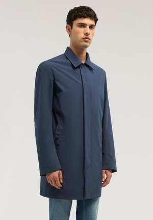 Uomo che indossa un cappotto blu navy fino al ginocchio con tasche con bottoni, in piedi davanti a uno sfondo semplice grigio chiaro.