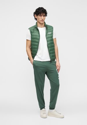 Zelena prešita telovnik čez belo majico, združena z zelenimi trenirkami. Oblečeno s svetlimi supergami, minimalističnega oblikovanja z logotipom.