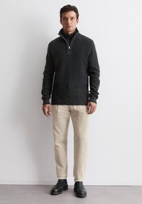 Dark gray knitted pullover with a high collar and half-zip, paired with light beige pants and black ankle boots on a plain background.