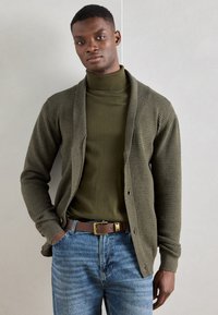 Olīvu zaļš kardigāns ar teksturētu dzijas dizainu, atvērta priekšpuse un ribotas piedurknes, uzvilkts virs atbilstoša turtleneck un ziliem džinsiem.
