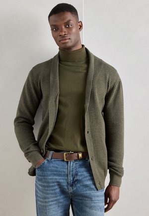 Olivgrüner Cardigan mit strukturiertem Strickdesign, offener Vorderseite und gerippten Bündchen, getragen über einem passenden Rollkragenpullover und blauen Jeans.