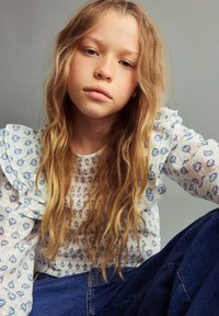 Blusa bianca a maniche lunghe con motivi floreali blu e polsini arricciati, abbinata a jeans blu scuro. Capelli lunghi, mossi e biondi chiari.