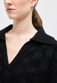 Femme portant un pull en V noir texturé avec un large col et une petite boucle d'oreille argentée, les cheveux attachés.