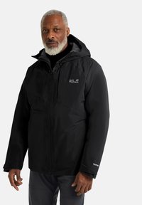 Giacca impermeabile nera di Jack Wolfskin con zip frontale, cappuccio e tessuto testurizzato; presenta dettagli con logo sul petto e sulla manica.